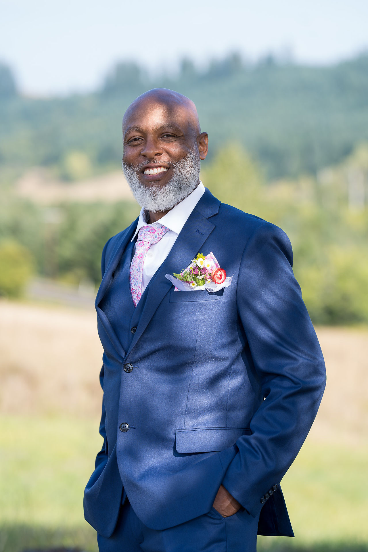 Smiling man in blue suit, pink tie, colorful pocket square outdoors.