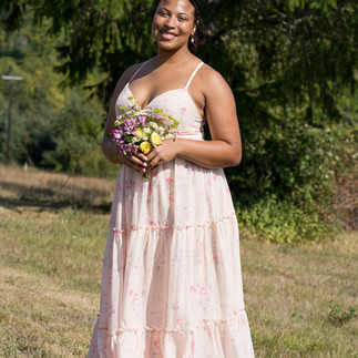 Smiling woman in a light pink dress holding a small flower bouquet outdoors.