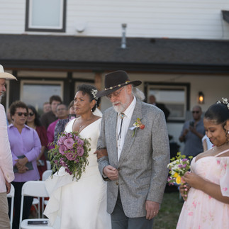 Bride with purple bouquet escorted by man at intimate farm wedding.
