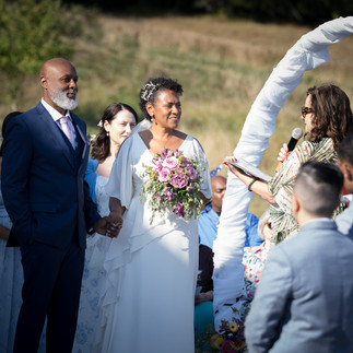Black couple holding hands, bride with bouquet, officiant at intimate farm wedding.