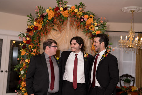 Three men in suits smile before a vibrant fall floral arch, White Salmon WA.