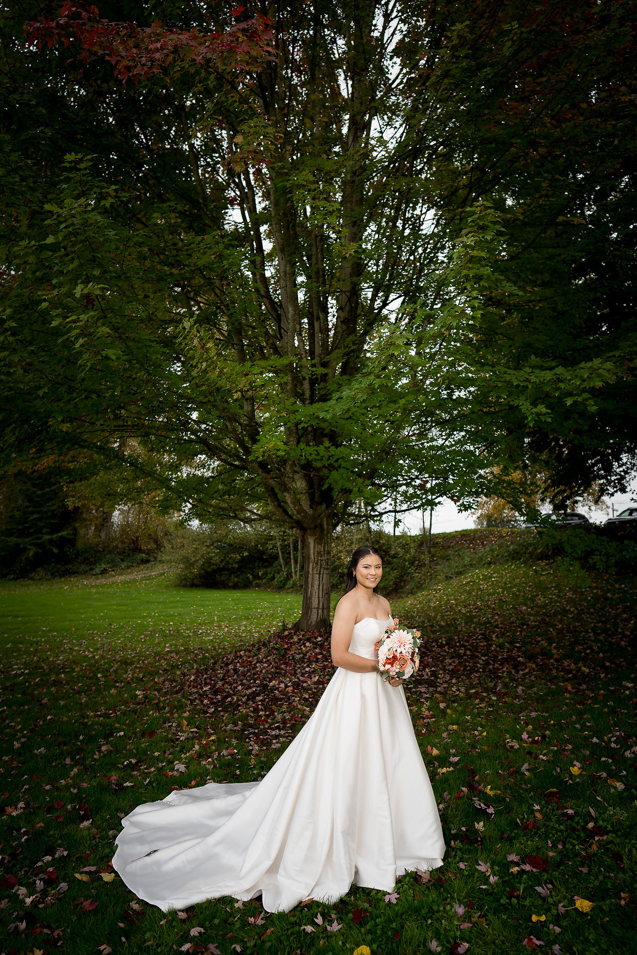 Elegant bride in white gown with bouquet under large green tree.