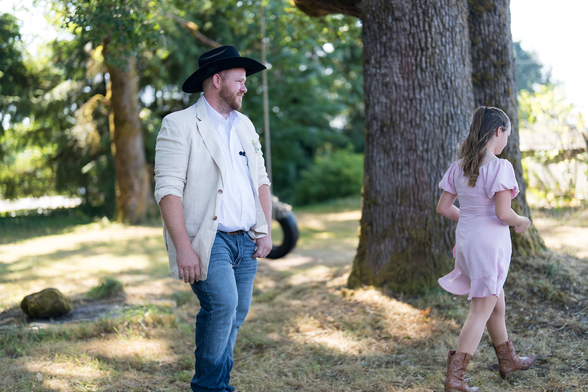 Man in cowboy hat, jacket, jeans in rustic outdoor setting, woman partially visible.