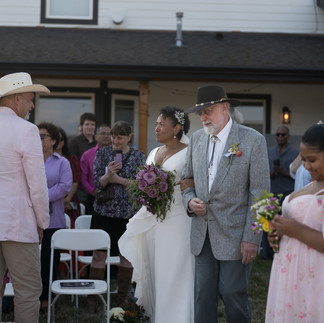 Bride escorted by man in hat at intimate outdoor farm wedding.