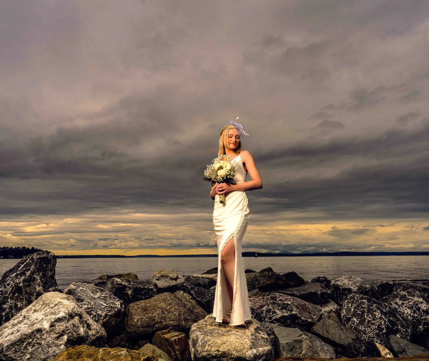 Woman in white wedding dress on rocks by water, cloudy sky, PNW wedding photographer.
