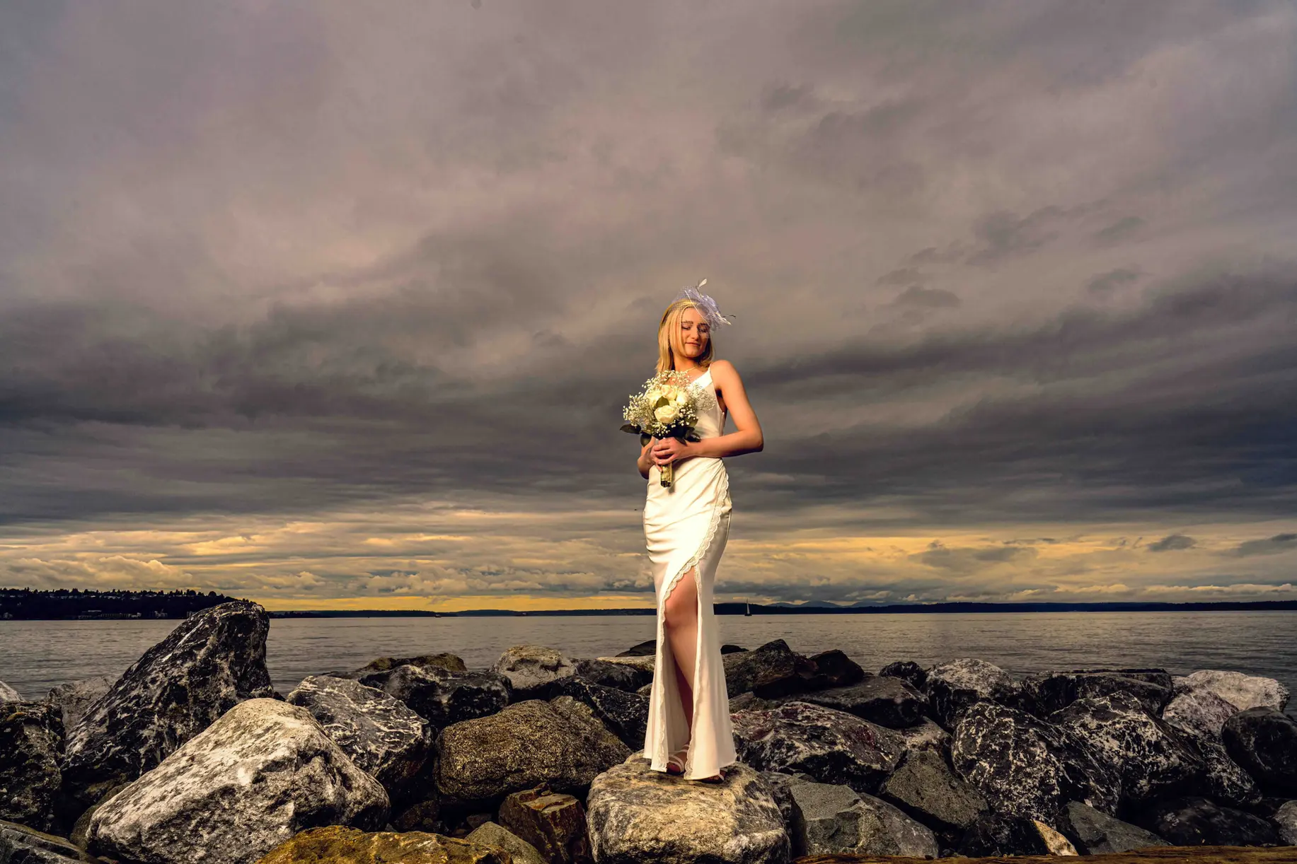 Woman in white wedding dress on rocks by water, cloudy sky, PNW wedding photographer.