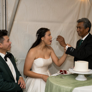 Bride in white dress fed cake by groom while another watches.