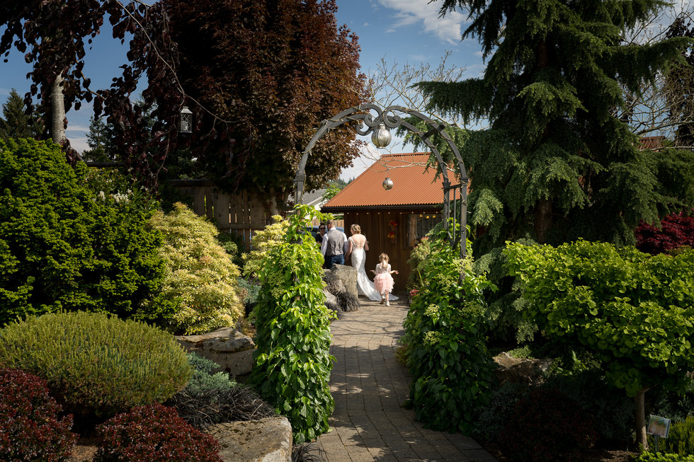Wedding party walks through arched garden path to outdoor venue.