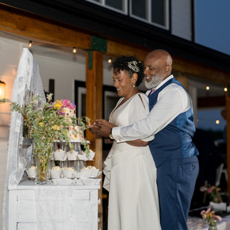 Joyful Black couple cutting tiered wedding cake and cupcakes outdoors.