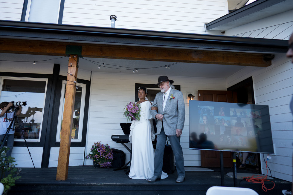 Bride and older man stroll on porch at intimate farm wedding.