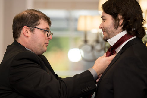 Man in glasses adjusts another man's red tie before a formal event.