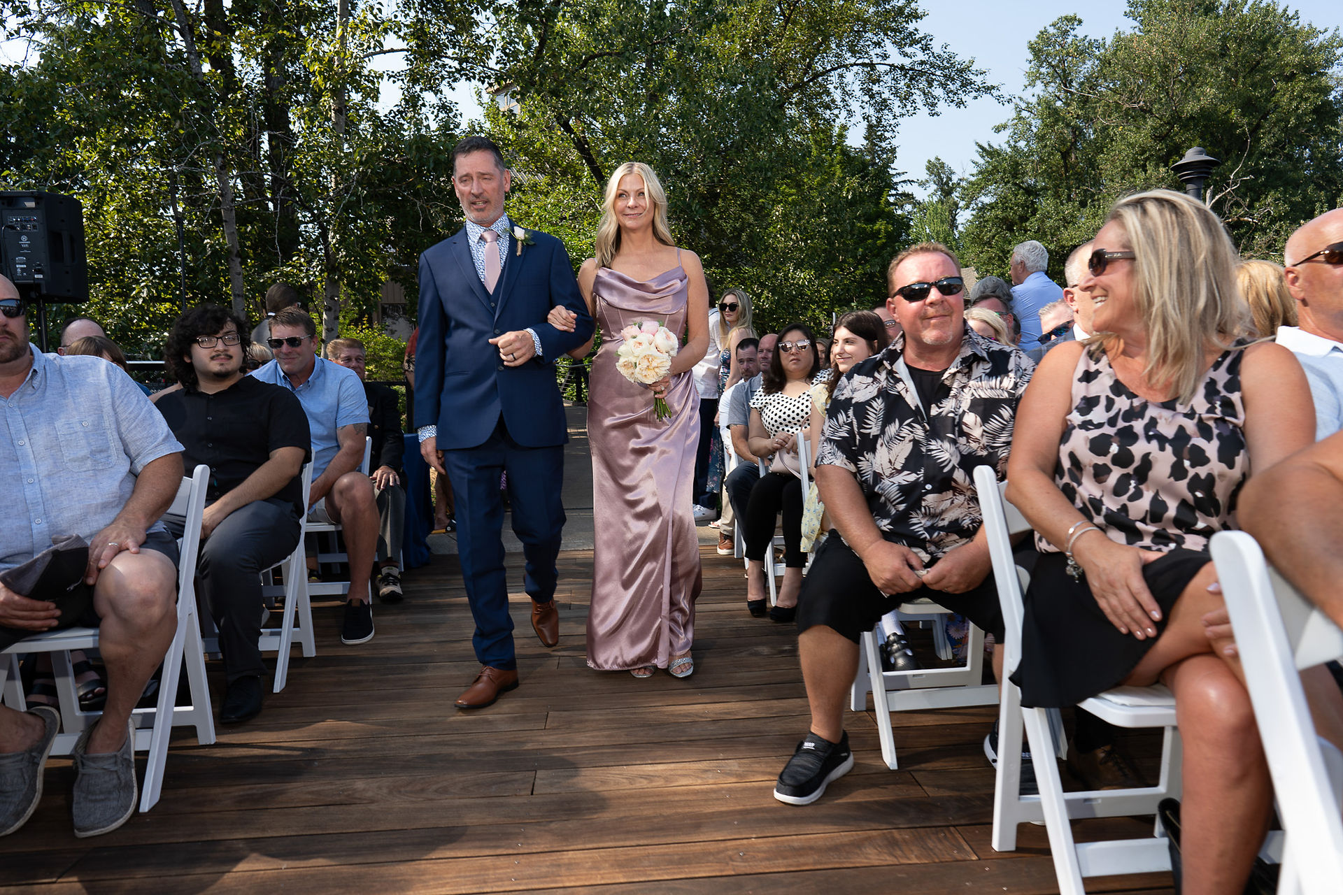 Man, woman in lavender dress walk wedding aisle, guests watch.