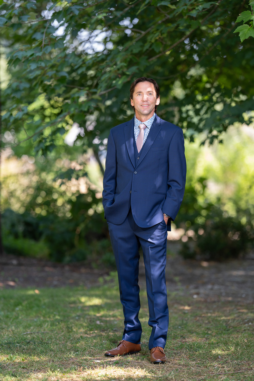 Man in blue suit smiles, standing outdoors among green trees.