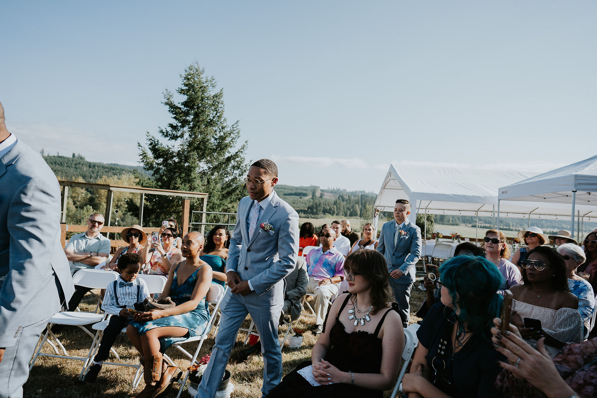 Groom in blue suit walks past guests at an outdoor farm wedding.