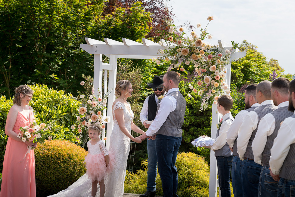 Bride, groom hold hands under floral arch at outdoor wedding ceremony.