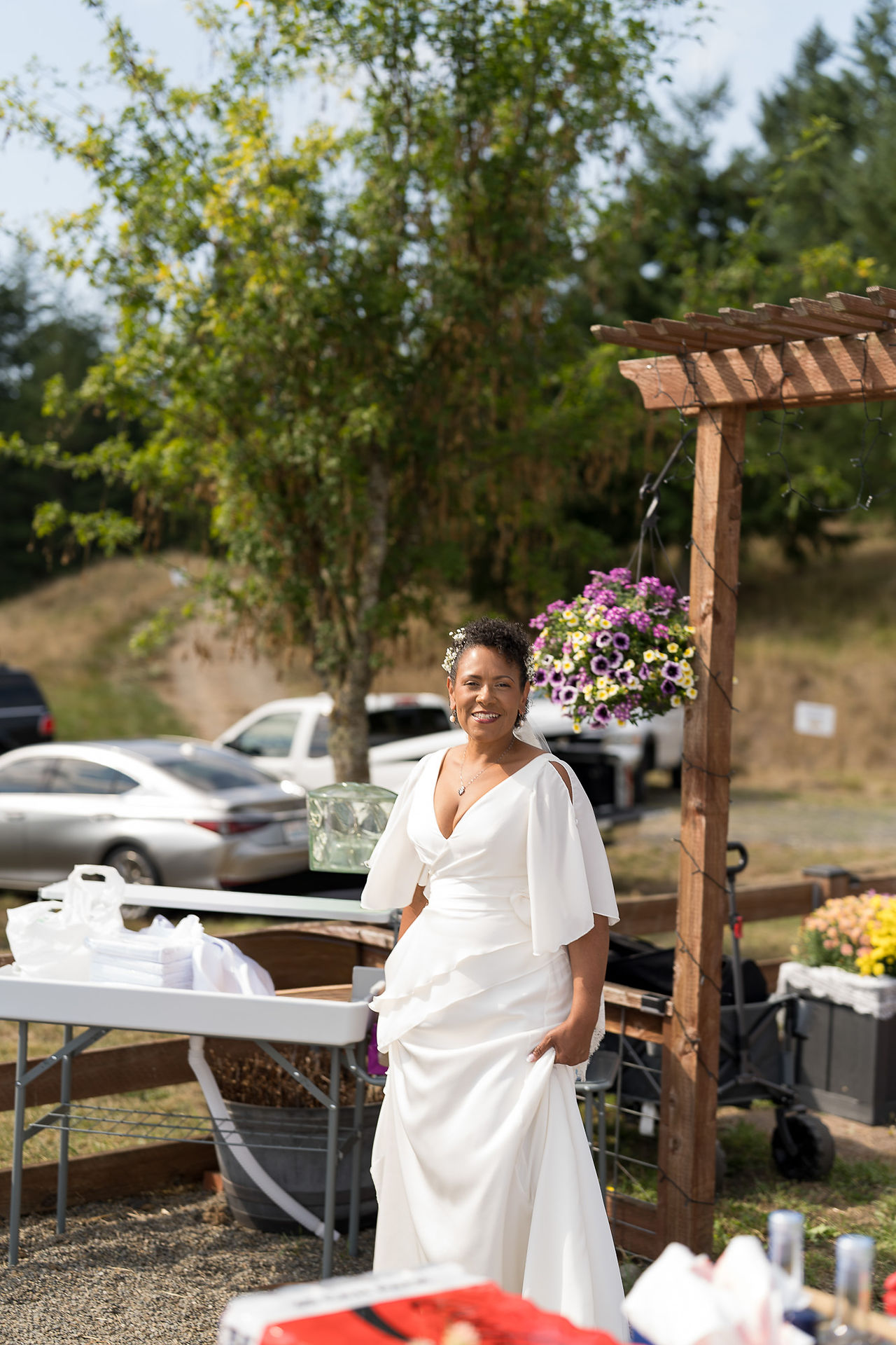 Smiling woman in white wedding dress standing outdoors near wooden arch.