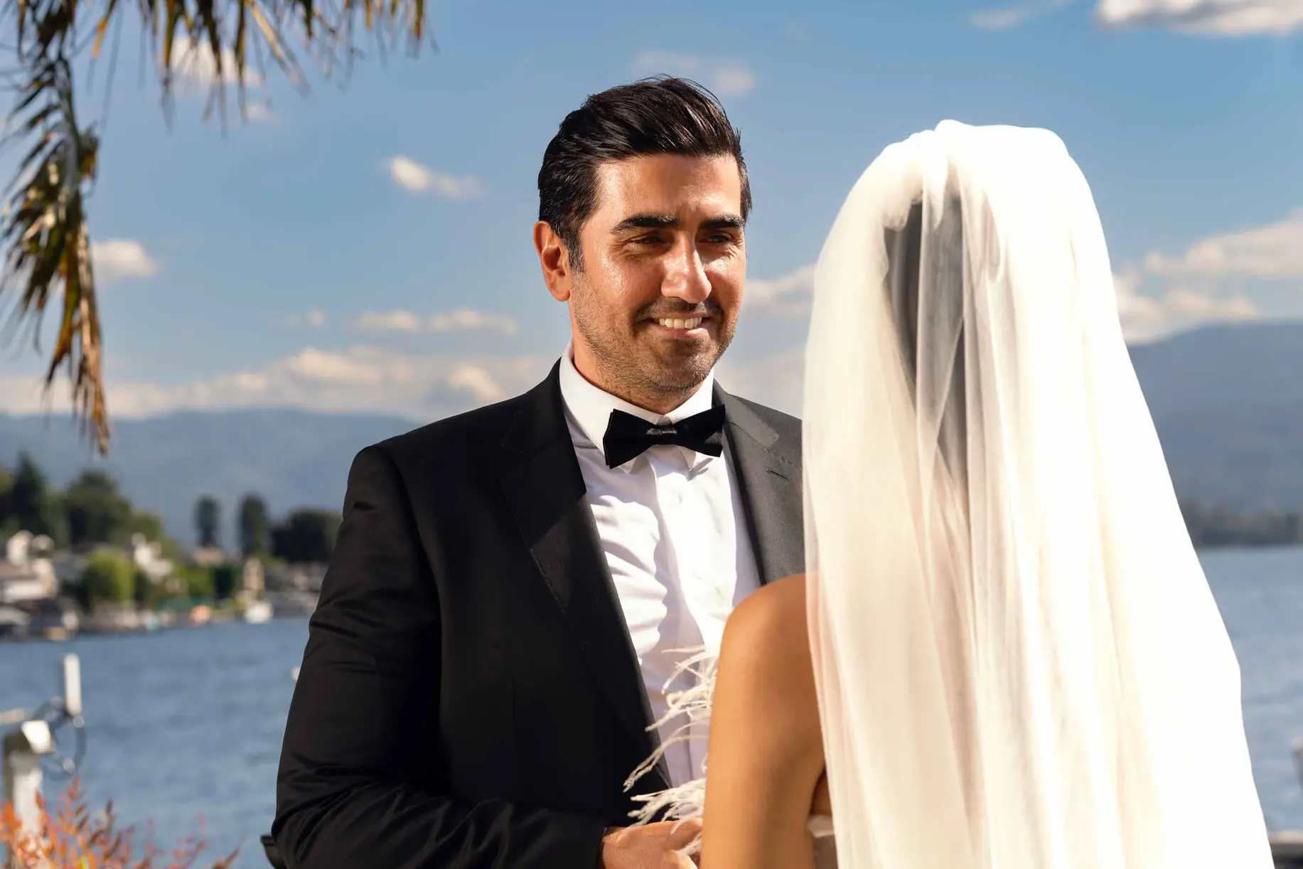 Groom smiling at bride during outdoor sunny wedding by lake.