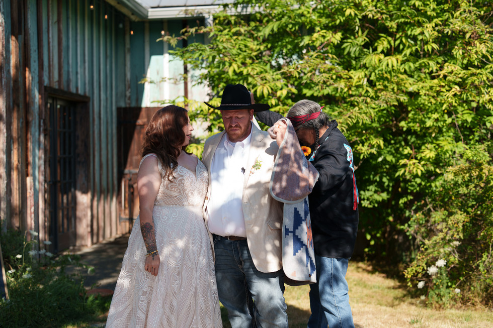 Bride watches groom as elder dresses him at outdoor wedding.