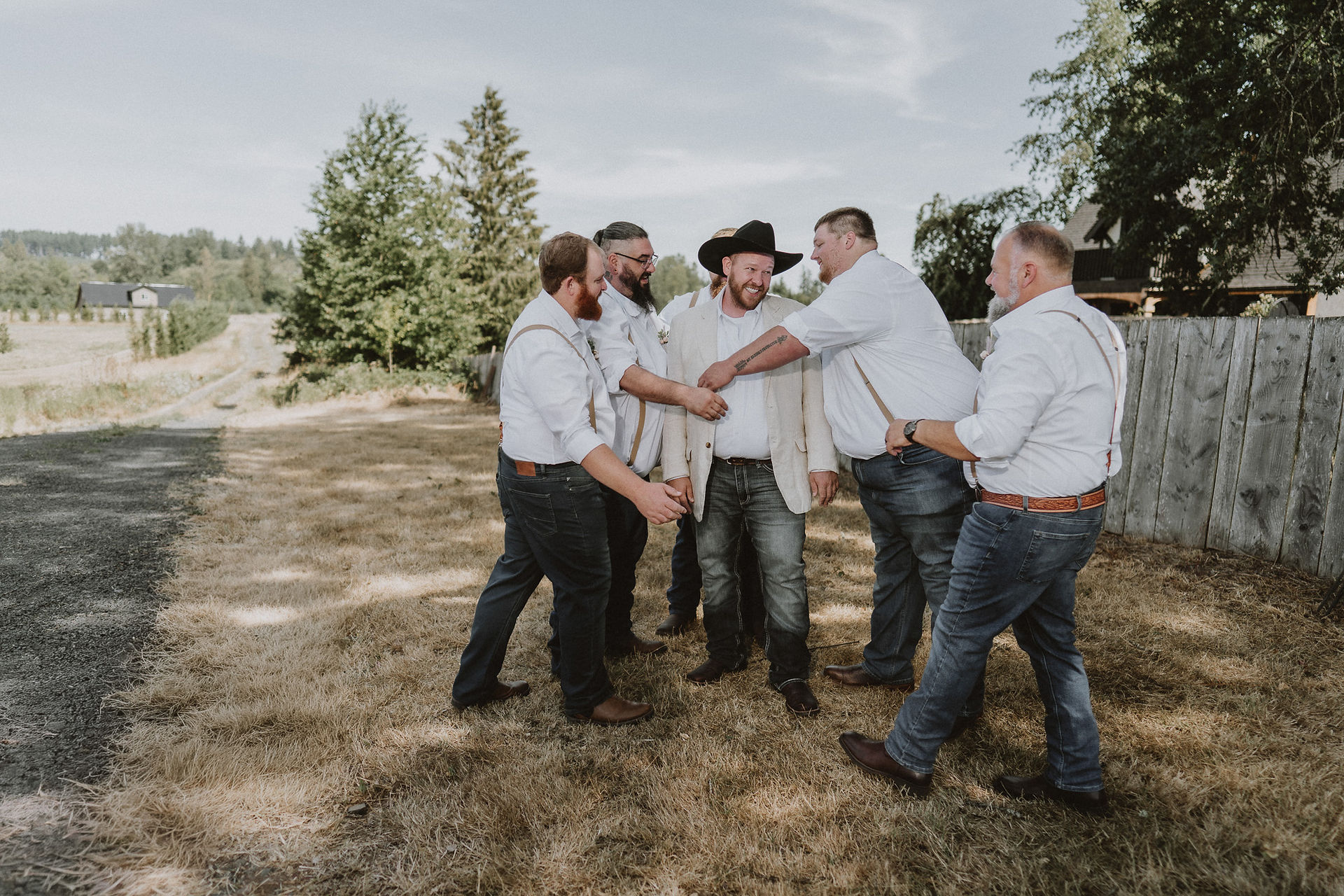 Groomsmen laughing and adjusting groom's jacket outdoors on wedding day.