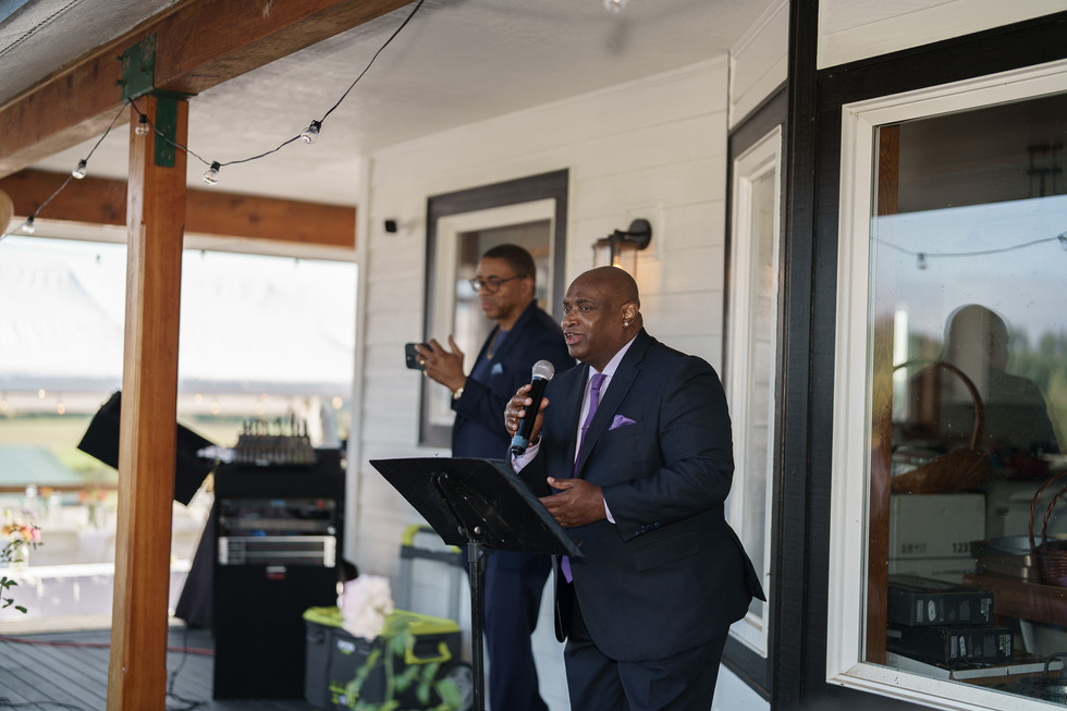 Man speaks into microphone while another claps during outdoor event.