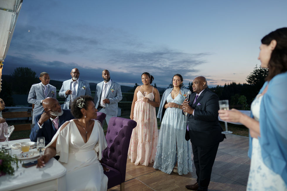 Wedding guests and bride listen as man toasts outdoors at dusk.
