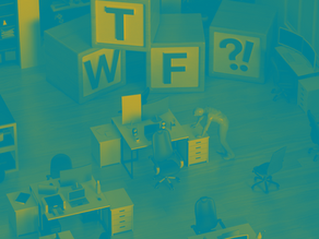 Office with large blocks spelling "T", "W", "F", and "?!" A person leans on a desk. Computers and chairs in the background.