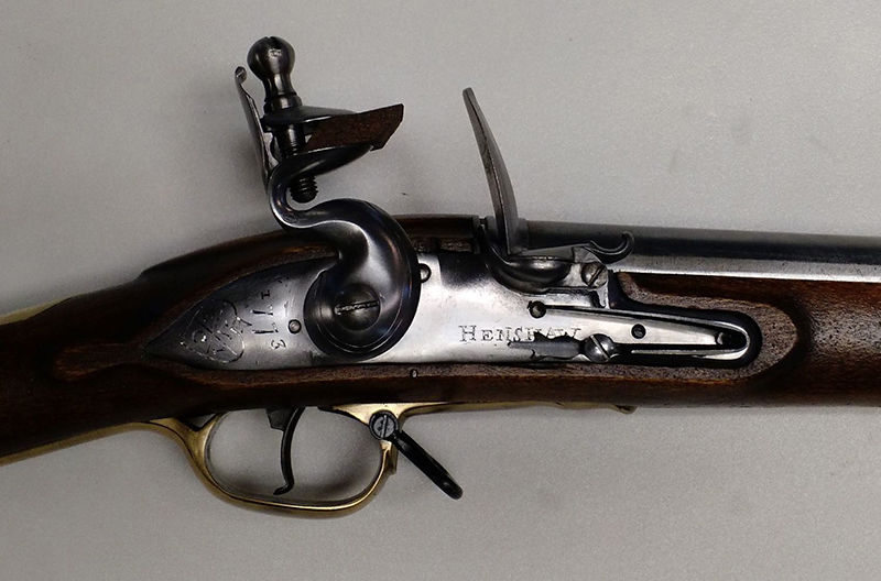 Close-up of a flintlock mechanism on a brown wooden musket. Metal components are engraved with text. Light gray background.