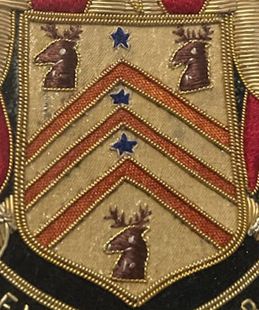 Embroidered crest with three red chevrons, three blue stars, and three brown deer heads on a gold background. Black and red edging.