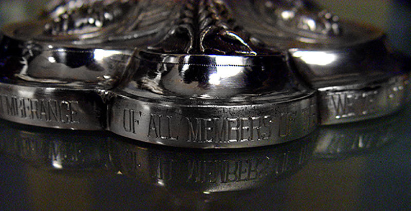 Close-up of a shiny silver trophy with engraved text: "OF ALL MEMBERS." The focus is on the reflective metal surface and intricate design.