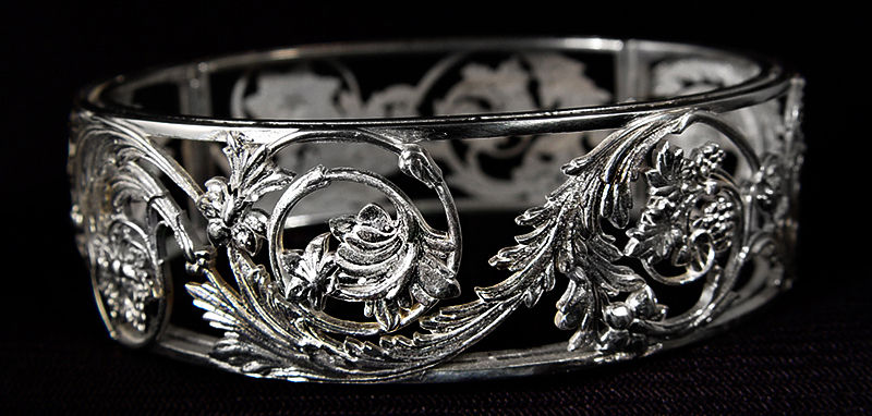 Ornate silver bracelet with floral designs on a dark background, showcasing intricate details and an elegant, antique style.
