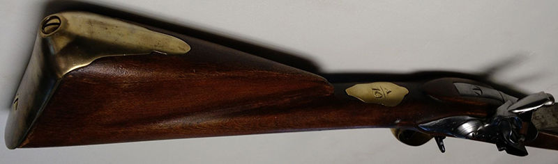 A close-up of a vintage rifle's wooden stock with brass fittings. The number "16" is etched on a brass plate. The background is plain gray.