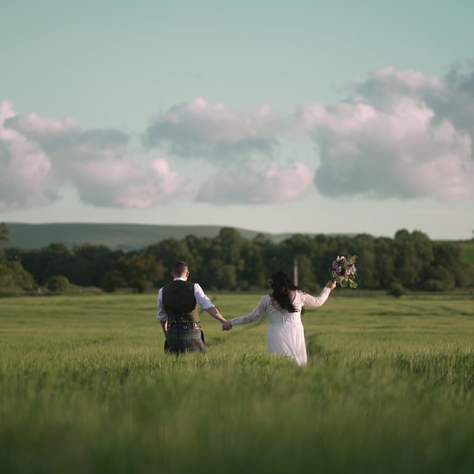 Newlywed couple walking hand in hand through a Scottish field, bride holding bouquet aloft