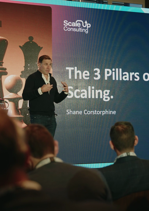 A man stands holding a microphone in front of a large screen. The text on the screen reads 'The 3 Pillars of Scaling.' There is an image of some pieces from a chessboard too. Two rows of people watch the presentation.
