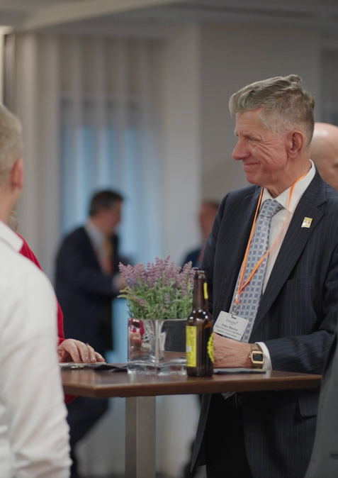 Three men and a woman chat around a poser table. They are laughing a smiling. The men are in shirts and ties. They are in a conference room and there are other people in the background.