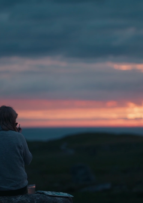 The sun is setting with beautiful colours of red and pink breaking through the clouds. A lone woman sits with her back facing us, she's painting what she sees.