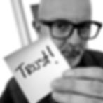Bald man with glasses holds a turquoise note reading "Trust!" in a room with framed art. Serious expression, casual attire.
