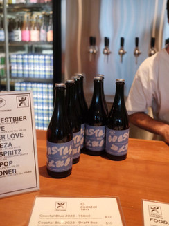 Homage Brewing bar with glass bottles of Coastal Blue and menus with Homage Brewing and Coastal Sun logos listing the taps and collaboration brew