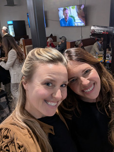 two women meet their heads together and smile at the camera while people talk in the background