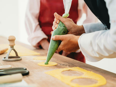 Hands piping green filling onto yellow pasta circles on a wooden table. Pasta cutter and tools visible. Warm, focused cooking atmosphere.