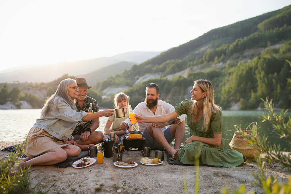 Smiling family group of luxury travelers picnic by a lake at sunset, raising drinks beside plates of food with green hills in the background.