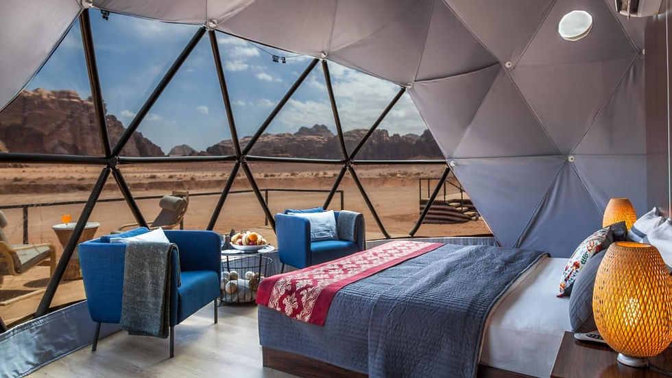 Inside a dome, a cozy room with blue chairs, a bed with red and gray bedding, and wicker lamps. Desert view through geometric windows.