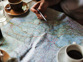 Hand holding a pen over an open road map at a table, beside coffee cups and a glass, suggesting travel planning