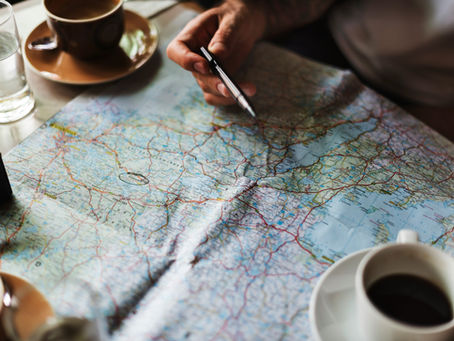 Hand holding a pen over an open road map at a table, beside coffee cups and a glass, suggesting travel planning
