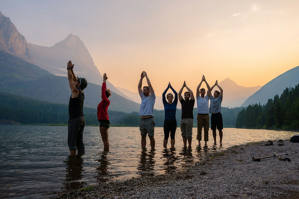 Seven people do yoga poses in a mountain lake at sunset, with calm water, pine forest, and glowing peaks.