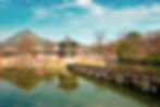Traditional Korean pavilion on a pond with pink blossoms, a red bridge, and a mountain in the background under a clear blue sky.