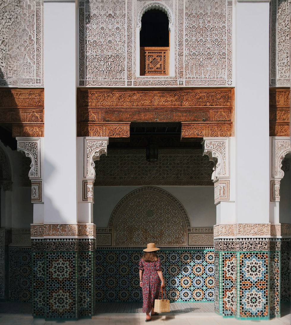 Woman in a straw hat and floral dress walks through an ornate Moroccan zellige tile work doorway carrying a straw bag.