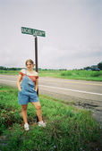 A young woman wearing short overalls and an orange off the shoulder top stands in front of a street sign reading "Rachel Carson Way". A field with a tractor is in the background.