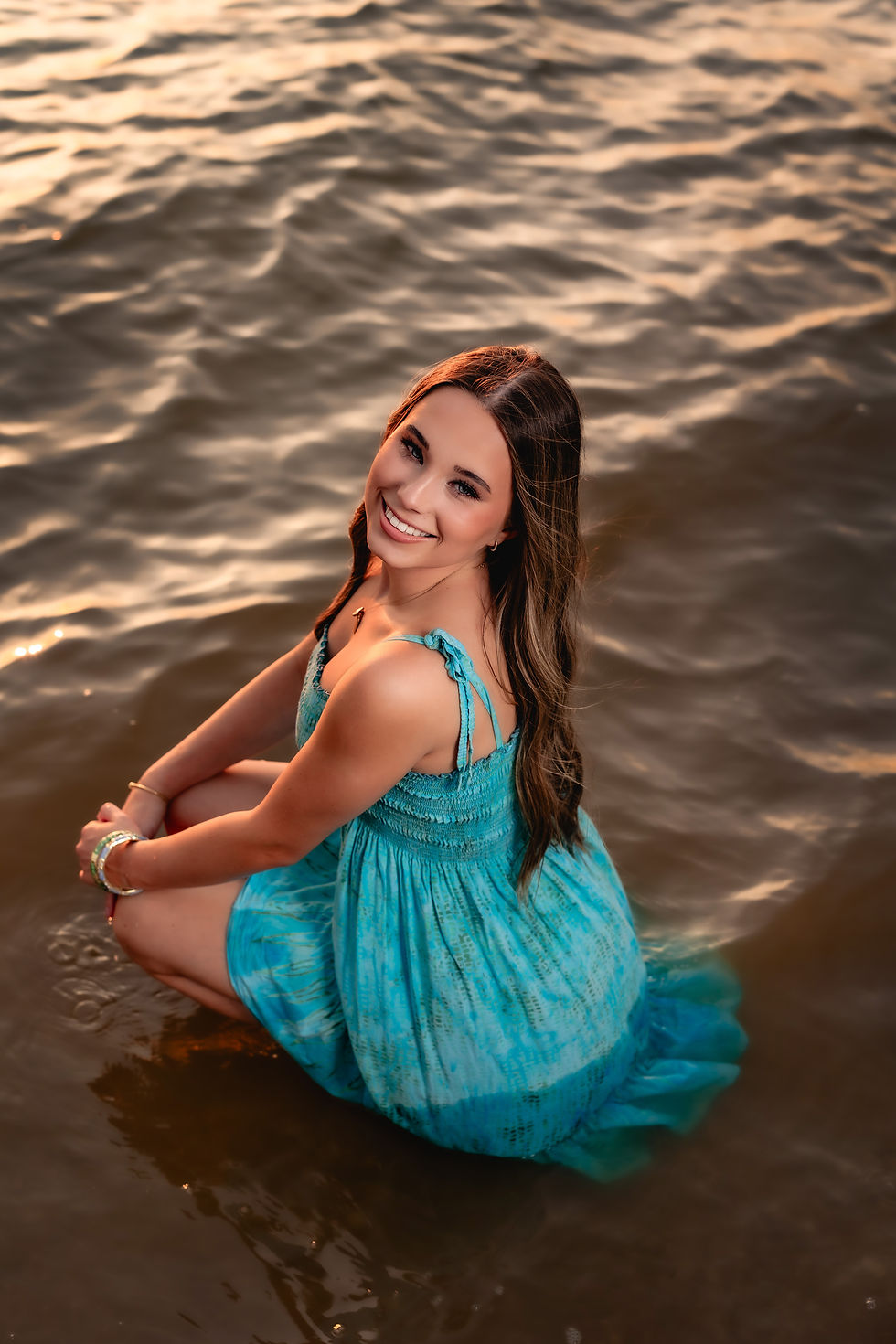 Smiling woman in a blue dress sitting in water during sunset. Gentle waves and warm colors create a serene and joyful ambiance.