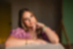 Woman in purple top, smiling softly with elbow on table, hand resting on chin. Wearing colorful bracelets, blurred neutral background.