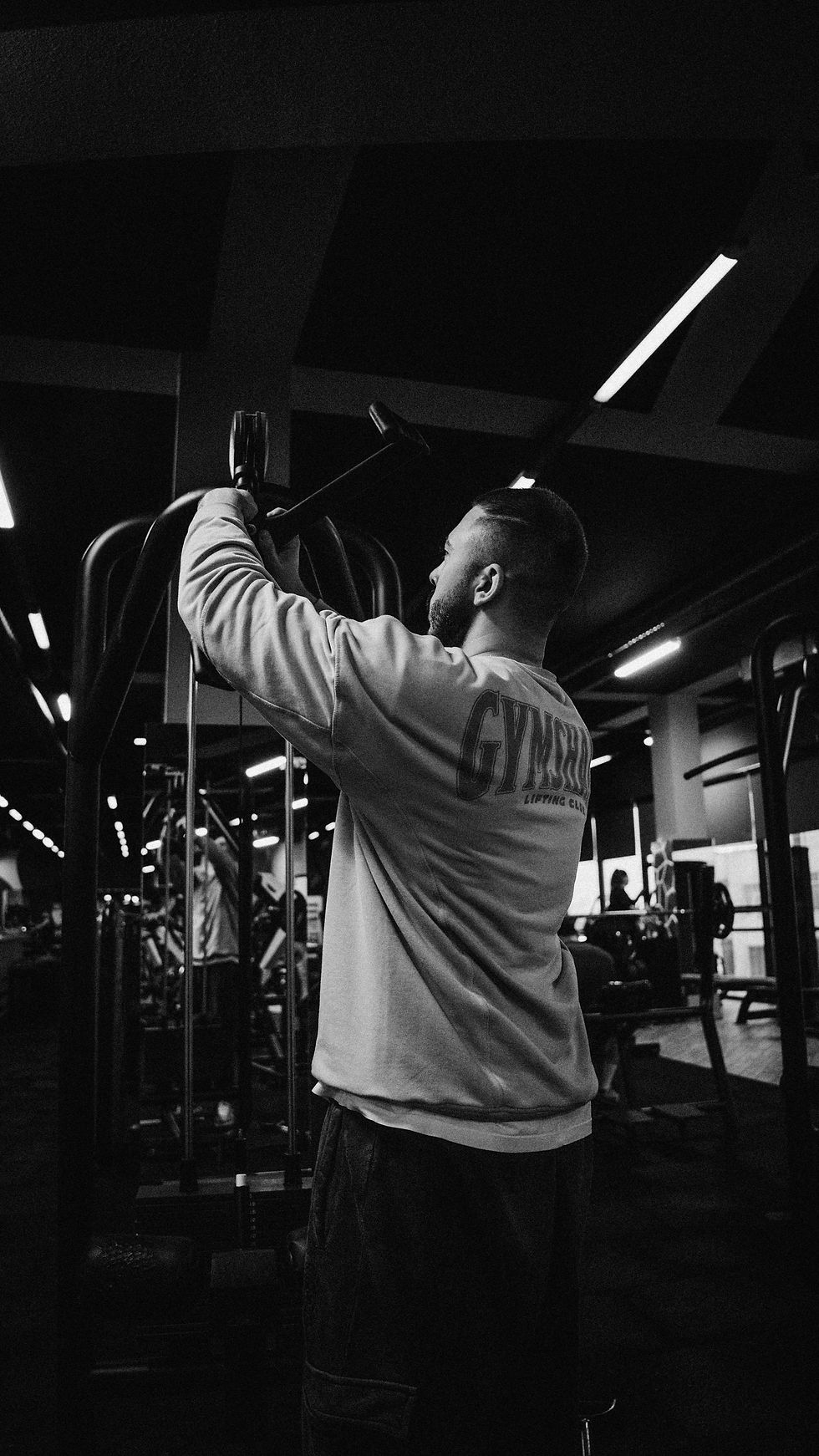 Man in a black-and-white gym setting adjusts a cable machine; sweatshirt reads GYMSTAR. Dark, focused workout mood.
